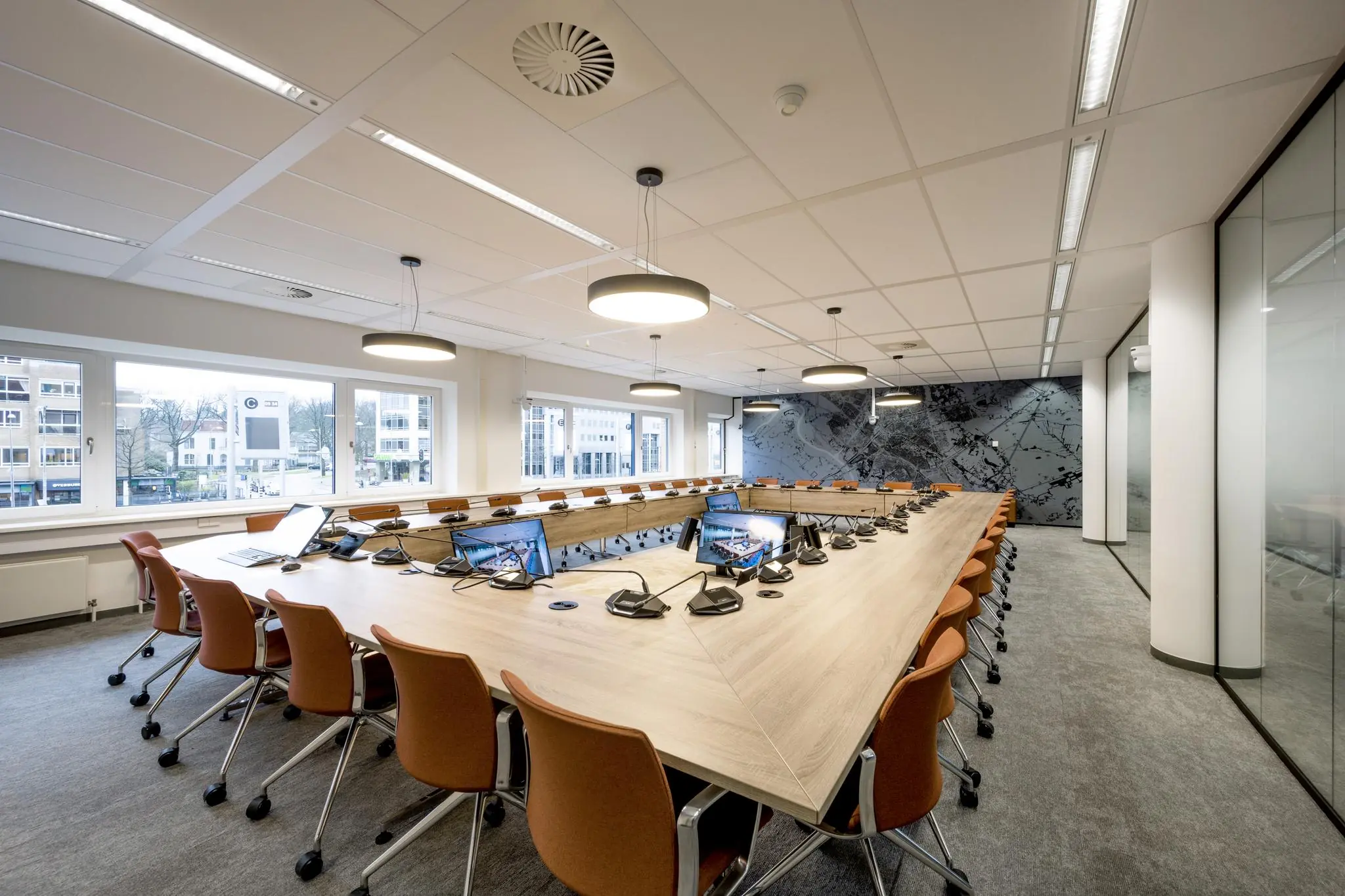 Vergaderruimte met een grote U-vormige tafel, microfoons, beeldschermen en oranje stoelen aan het Stationsplein.