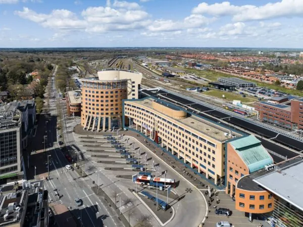 Luchtfoto van het Stationsplein in Apeldoorn met prominente ronde gebouwen, busperrons en het treinspoor op de achtergrond.