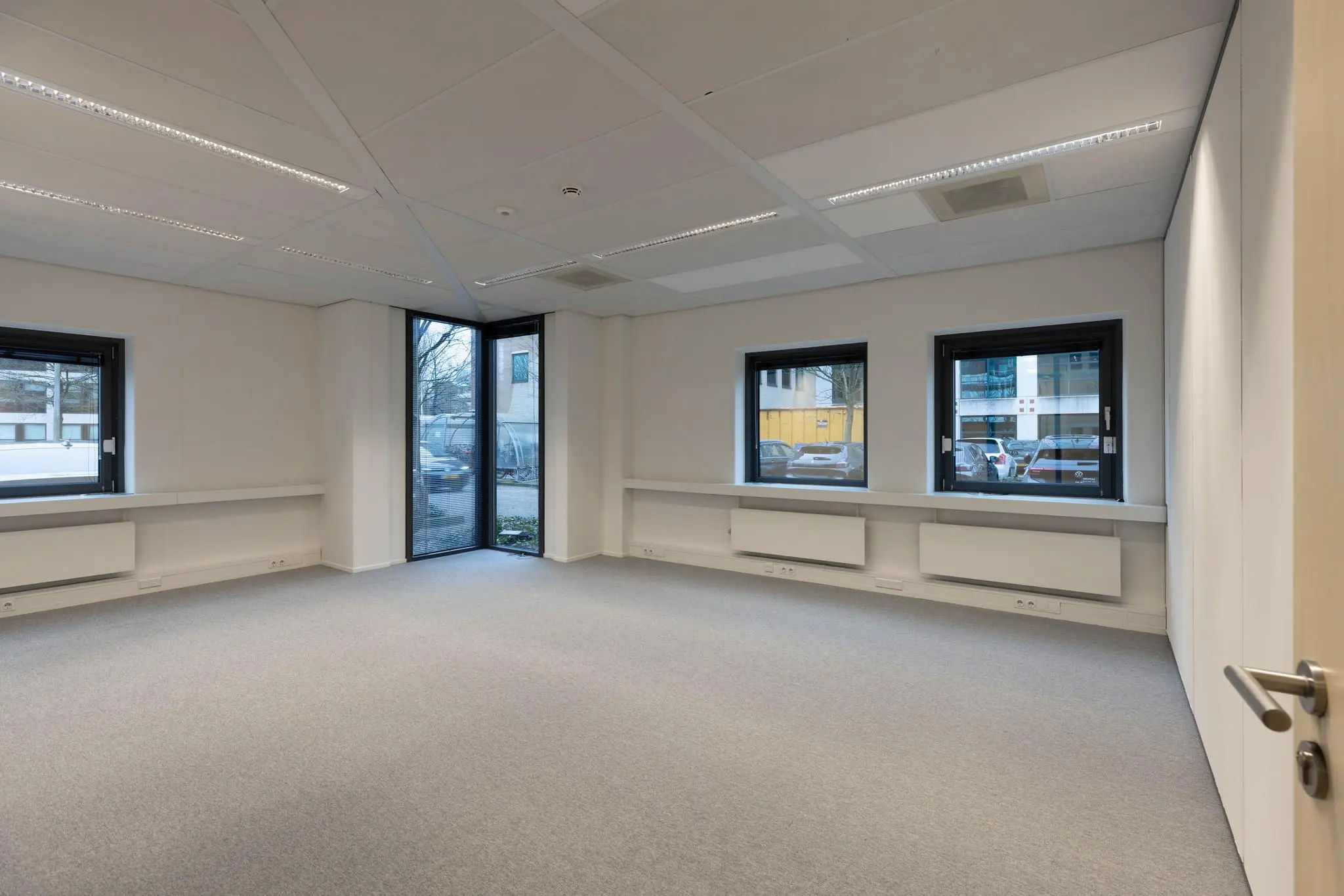 Leeg kantoorruimte met grijze vloerbedekking, witte muren en grote ramen met uitzicht op een parkeerterrein aan de Marathon.