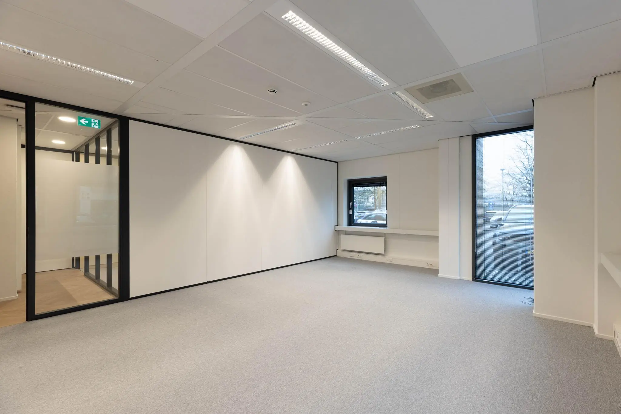 Leeg modern kantoor met witte muren, grijs tapijt en grote ramen met uitzicht op geparkeerde auto's.