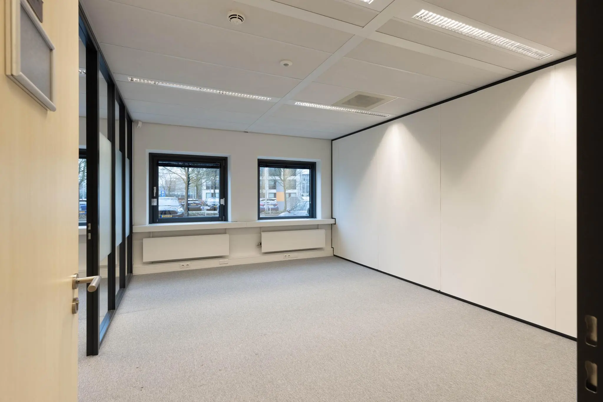 Lichte, lege kantoorruimte met drie ramen, witte muren en grijs tapijt in gebouw Marathon.