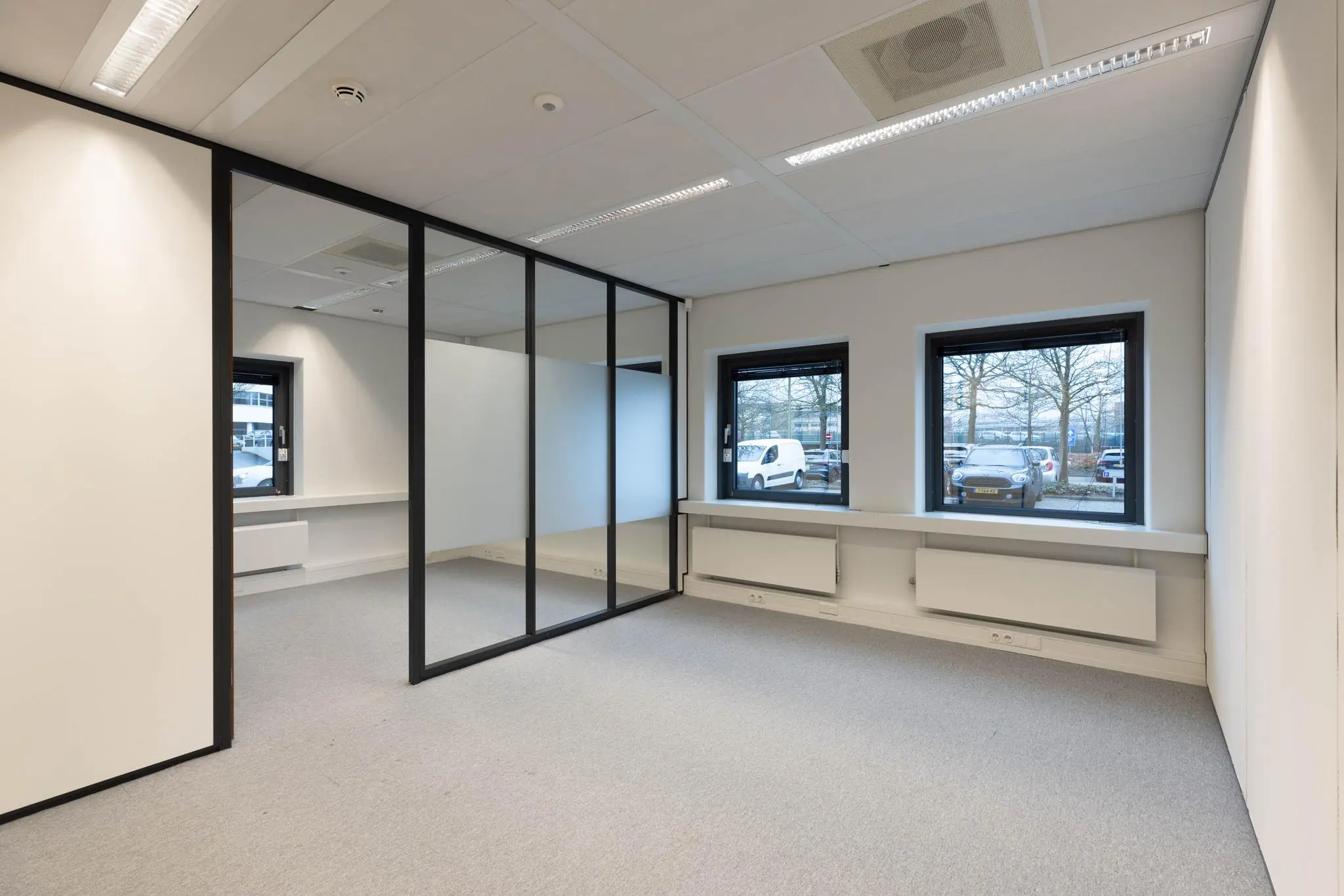 Licht en leeg kantoorpand met grijze vloerbedekking, glazen tussenwanden en uitzicht op een parkeerplaats.