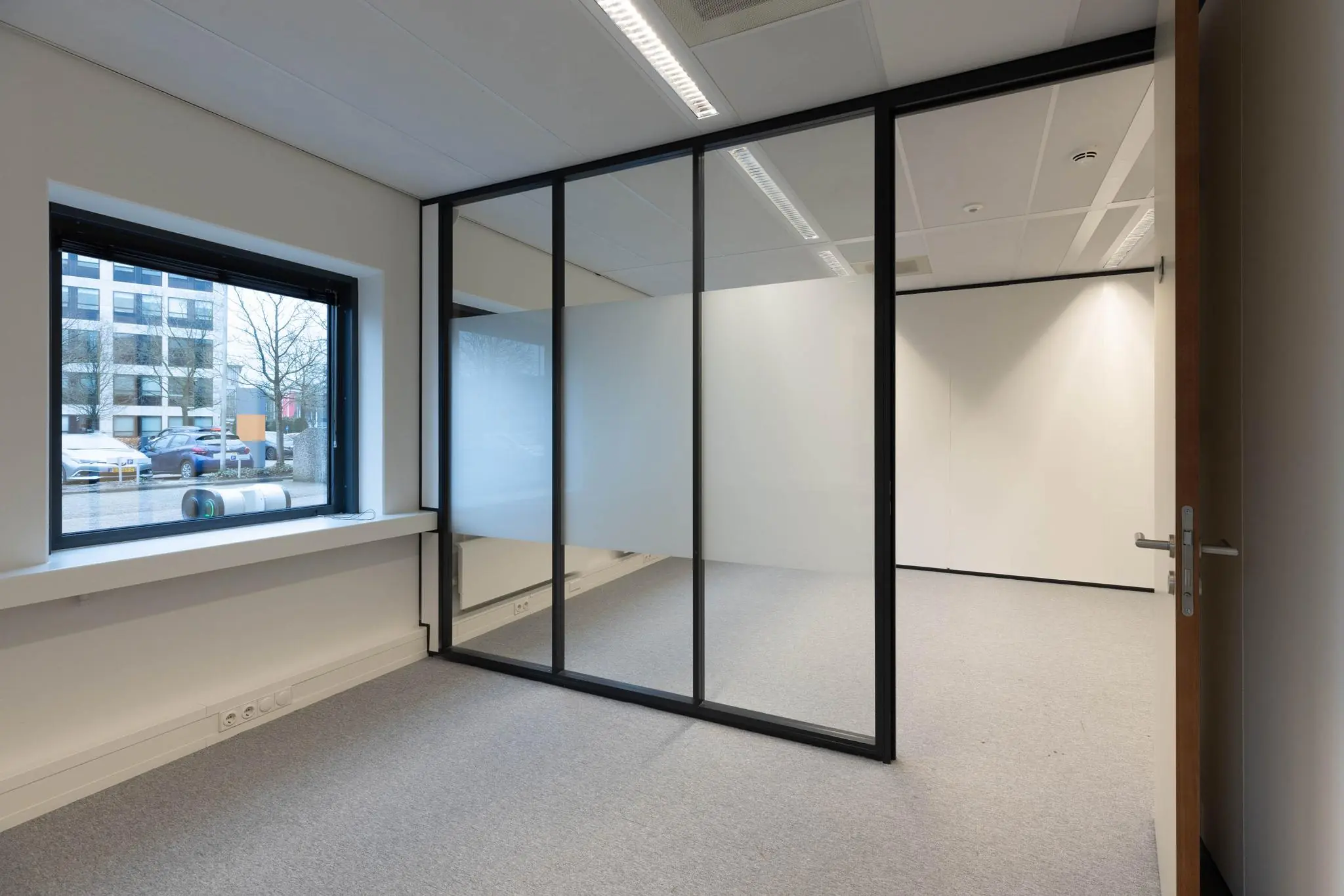 Leeg kantoor met glazen scheidingswand, grijze vloerbedekking en zicht op parkeerplaats buiten.