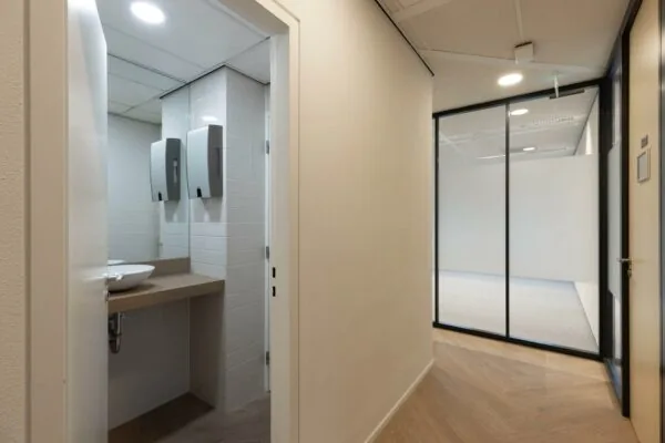 Gang in een modern kantoorpand met zicht op een toiletruimte en glazen scheidingswand naar een lege kantoorruimte.