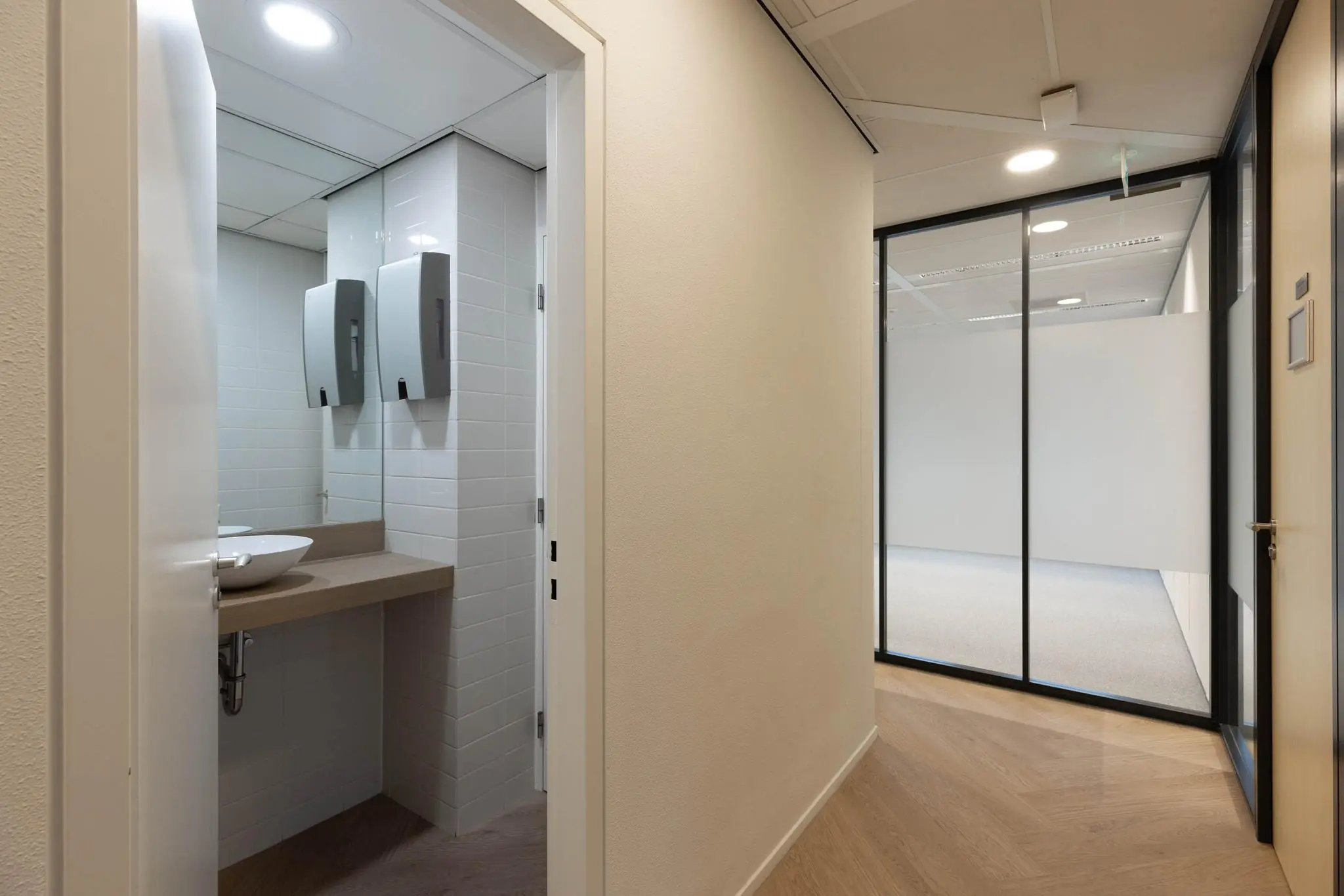 Gang in een modern kantoorpand met zicht op een toiletruimte en glazen scheidingswand naar een lege kantoorruimte.
