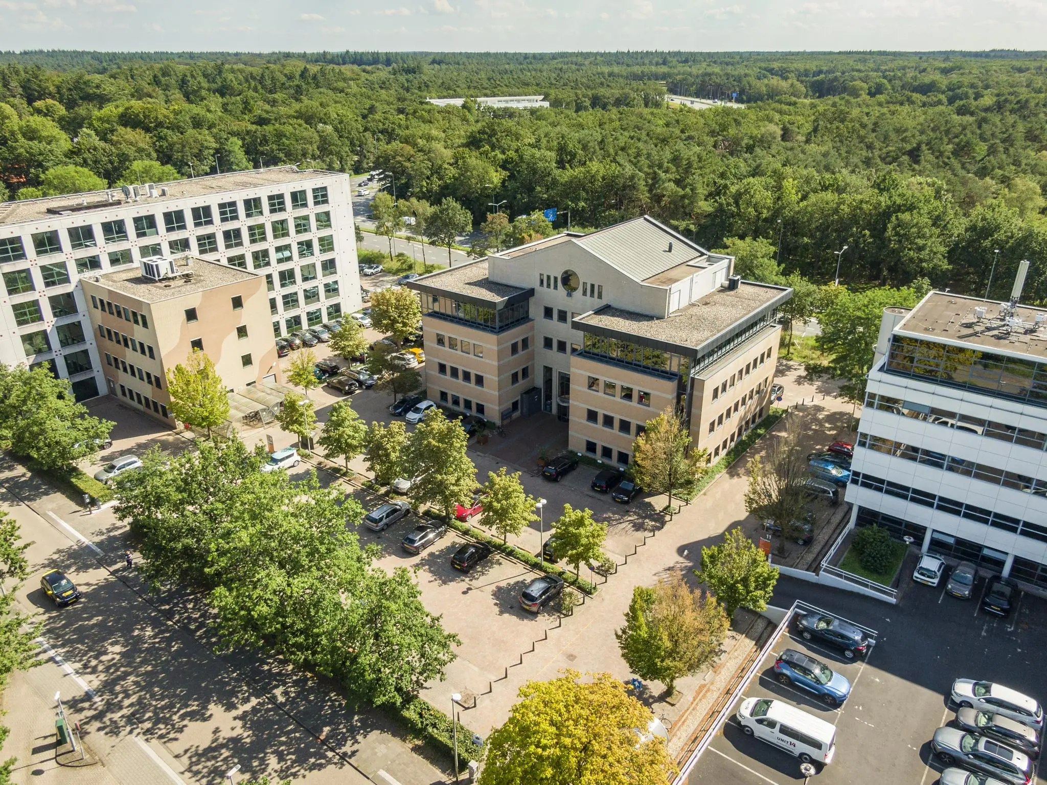 Luchtfoto van het kantoorgebouw Marathon in een groene, bosrijke omgeving met omliggende parkeerplaatsen en naastgelegen kantoren.