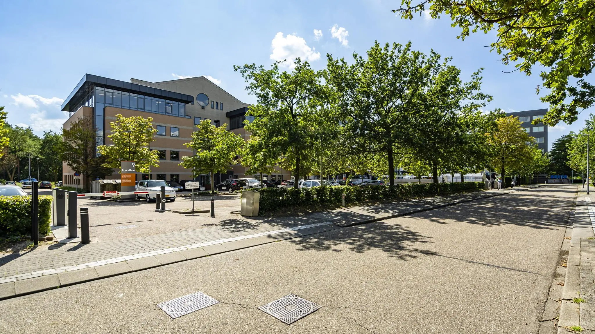 Kantoorgebouw Marathon 9 met parkeerplaats, omgeven door bomen aan een rustige straat op een zonnige dag.