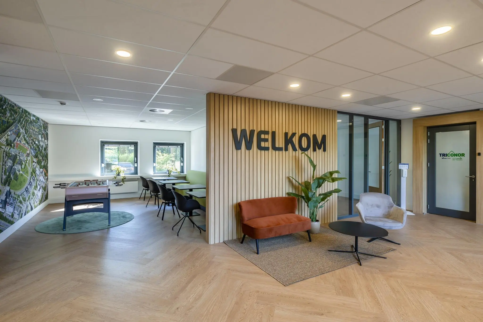 Moderne kantoorontvangstruimte met zitgedeelte, speeltafel en de tekst "WELKOM" op een houten wand.