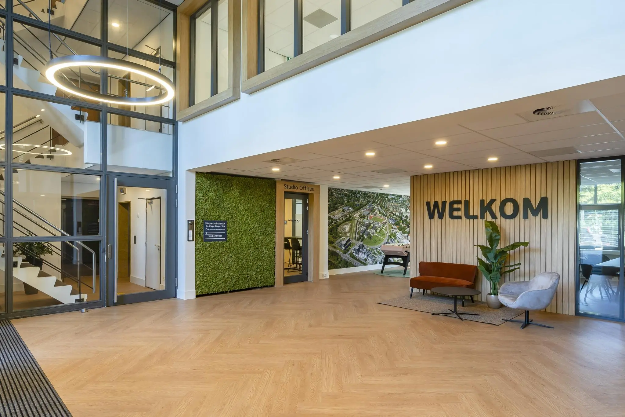 Moderne ontvangsthal met houten vloer, groene moswand en een groot "WELKOM"-bord aan de muur.