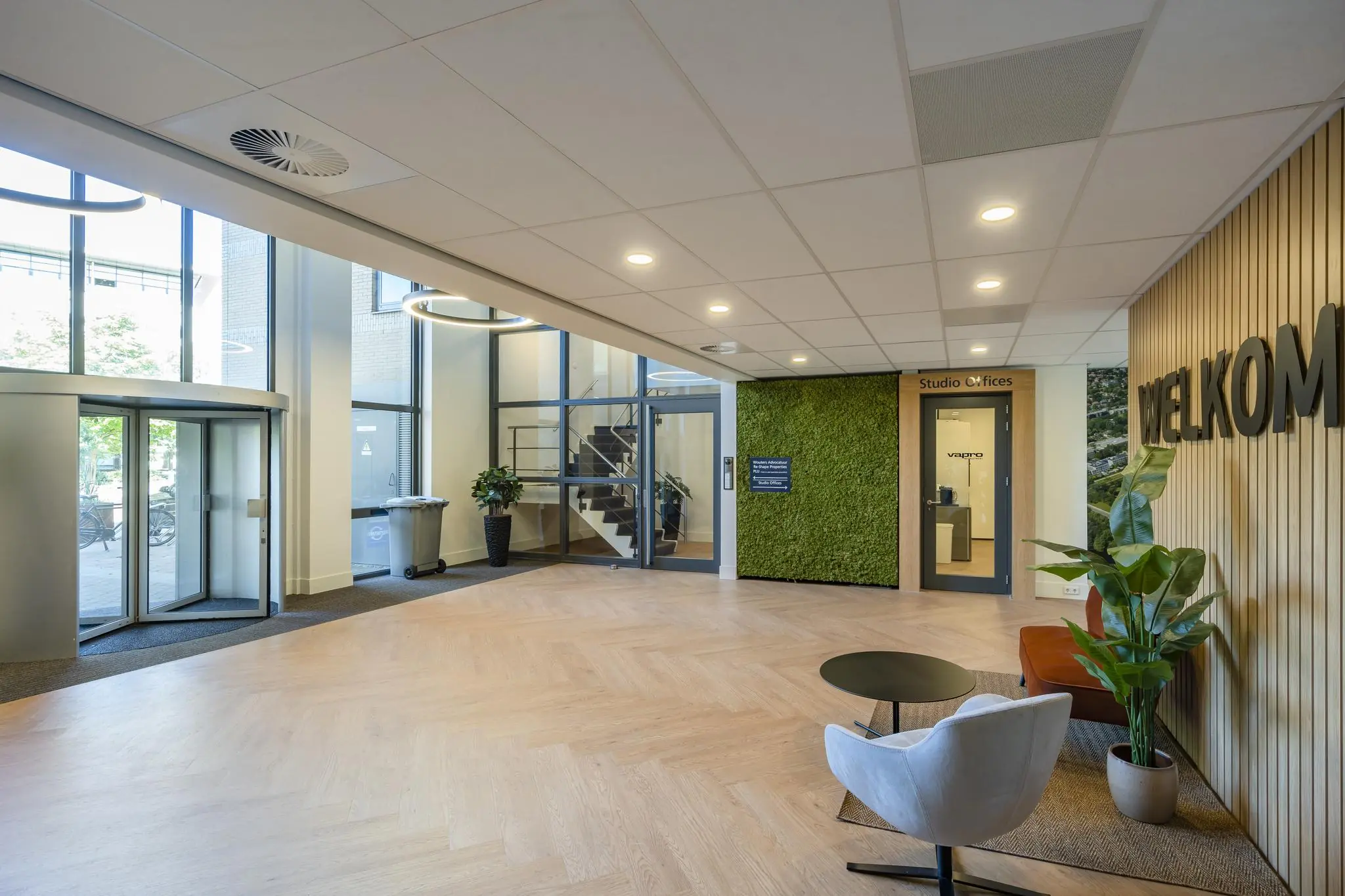 Moderne ontvangsthal van een kantoorgebouw met zithoek, groenemuur, en de tekst "WELKOM" op een houten wand.