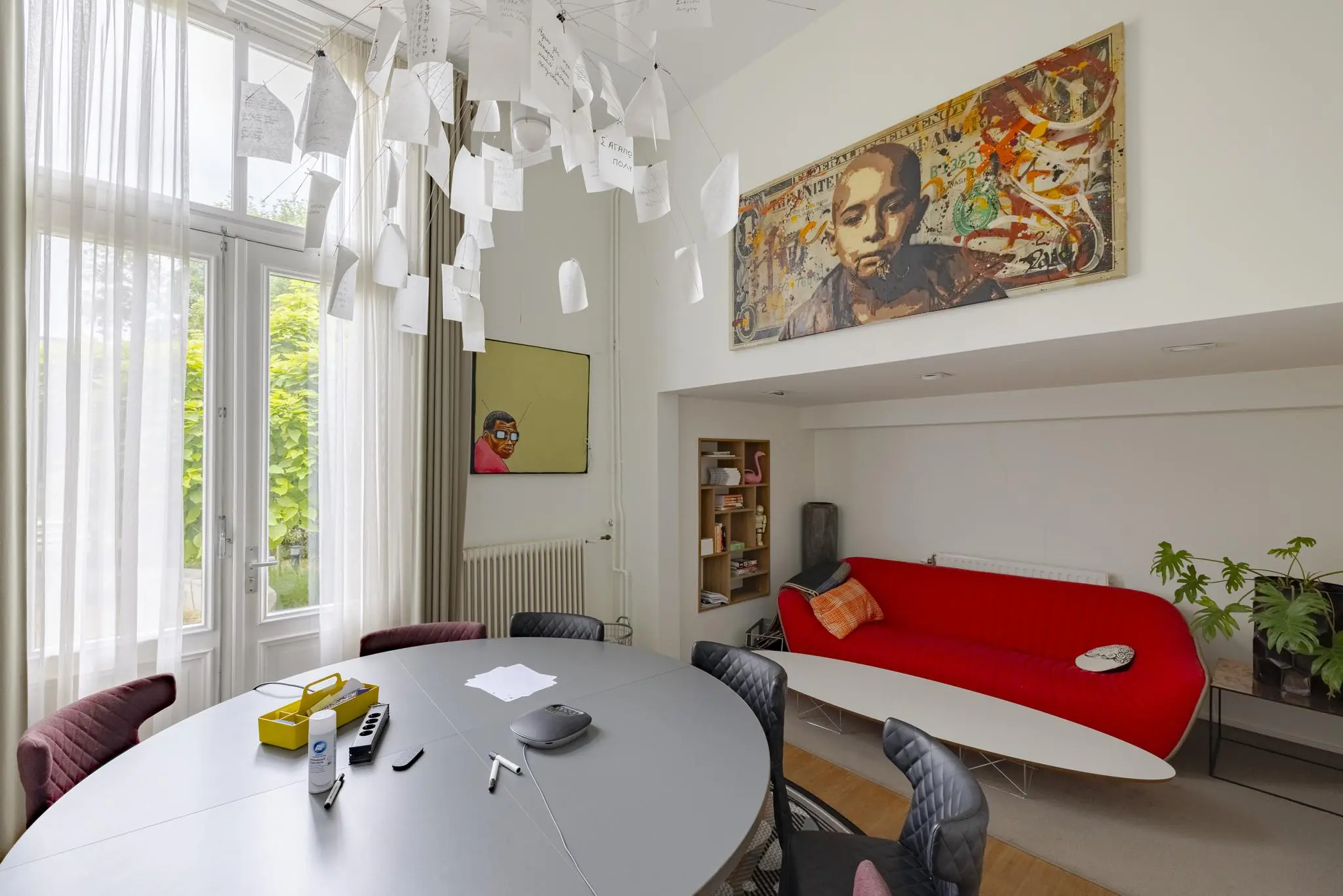 Vergaderruimte met ronde tafel, rode designbank, kunst aan de muur en een creatieve hanglamp met papieren.