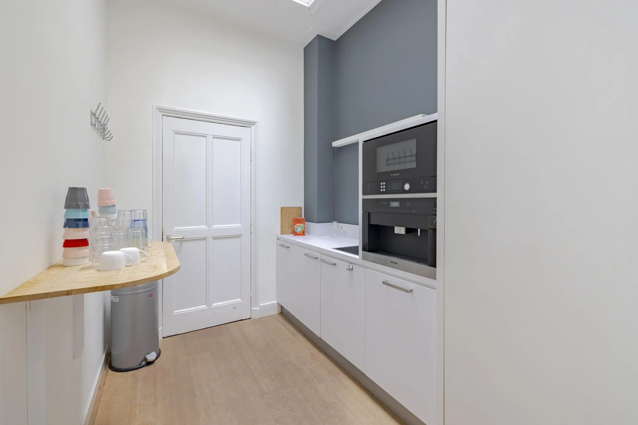 Kleine pantry met ingebouwde koffiemachine, aanrechtblad en stapels bekers op een smalle houten tafel.