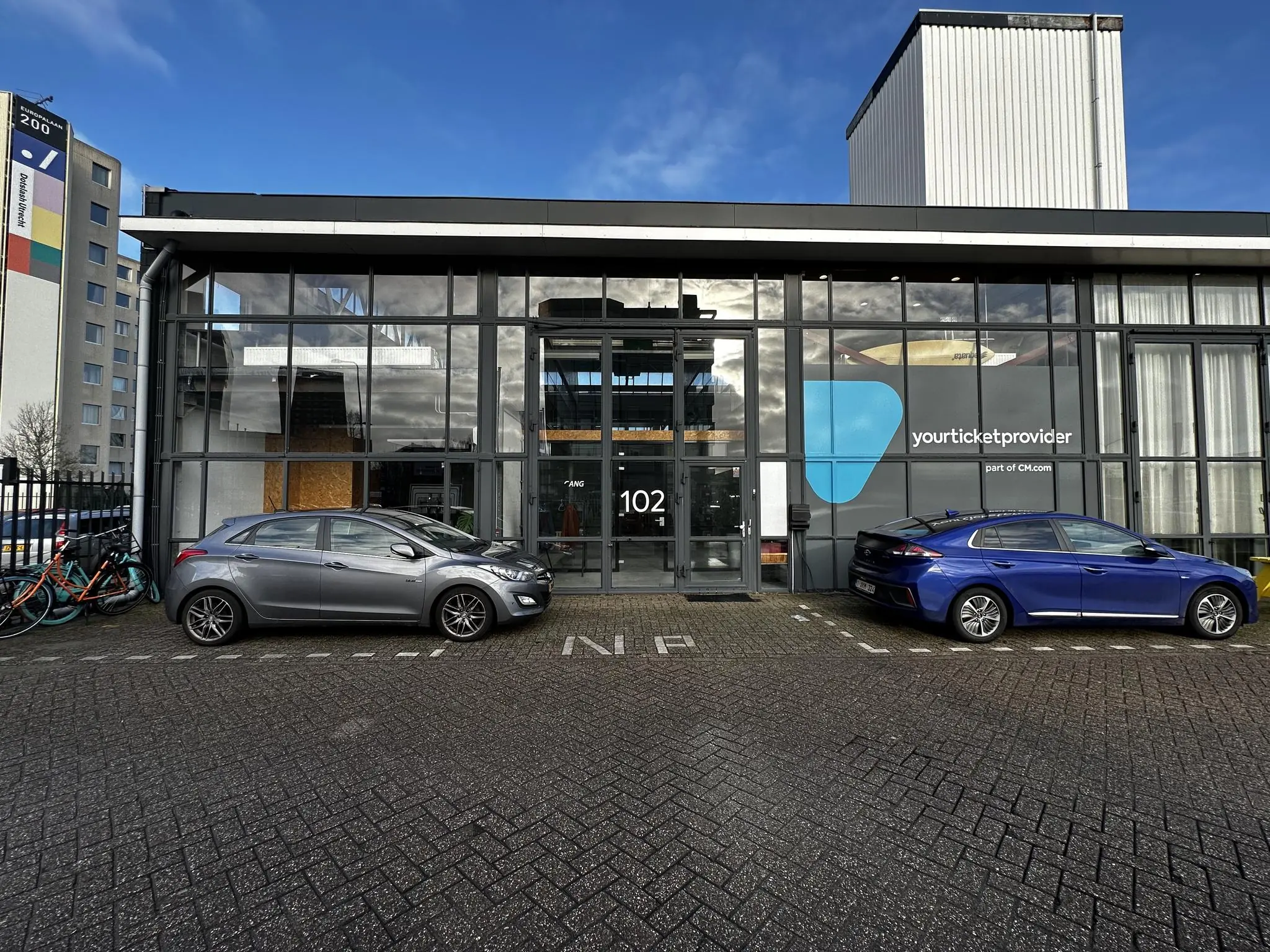 Vooraanzicht van het kantoorpand van Yourticketprovider aan de Eendrachtlaan 102, met geparkeerde auto’s en fietsen.