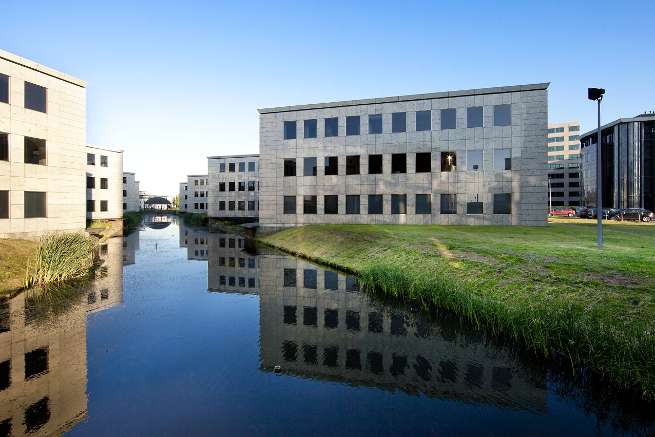 Kantoorgebouwen aan de Rivium 1e Straat weerspiegelen in het water van een aangrenzende gracht op een zonnige dag.