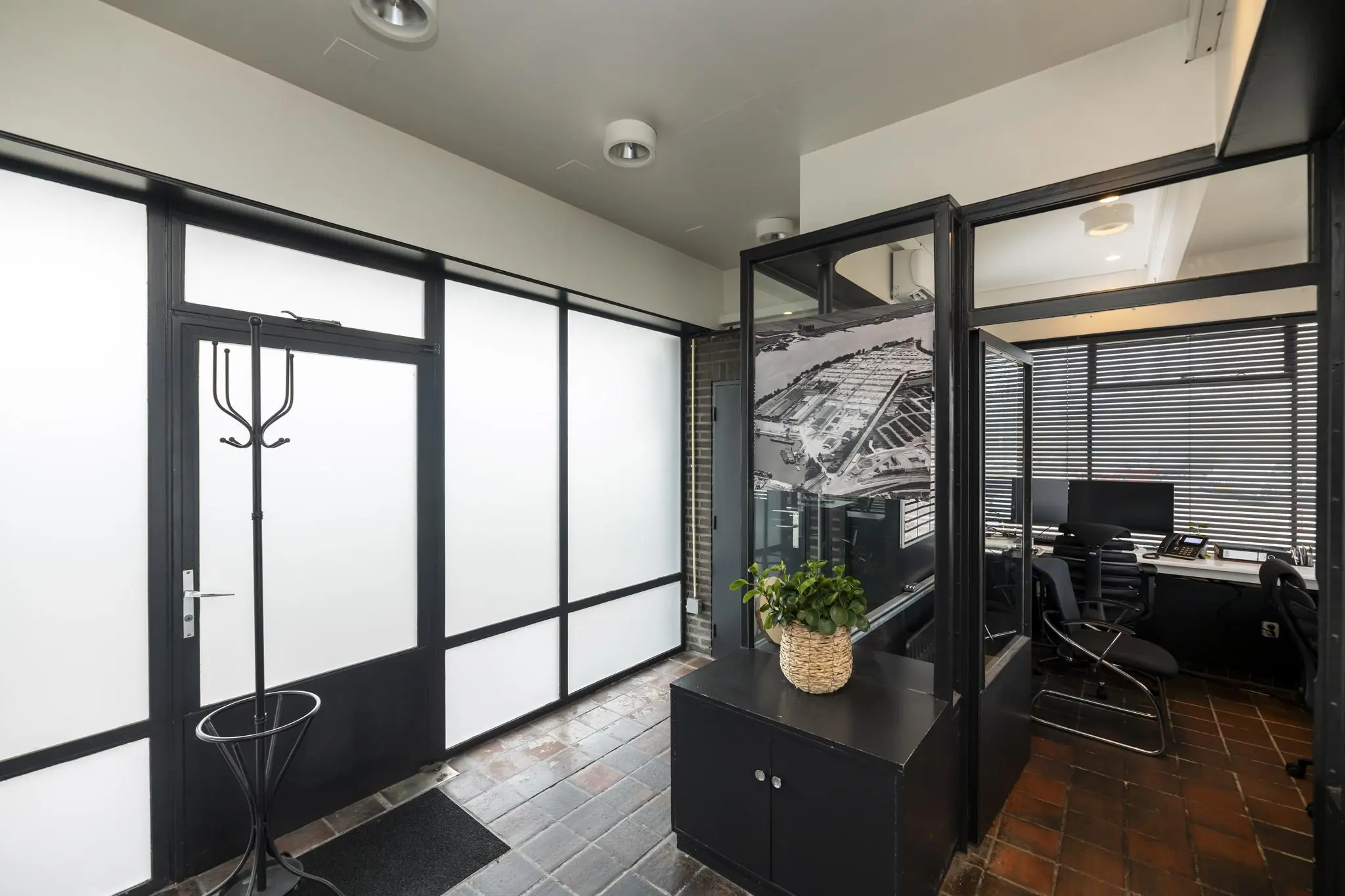 Moderne kantoorentree met zwarte raamkozijnen, glazen wanden en zicht op een werkruimte met bureaus en computers.