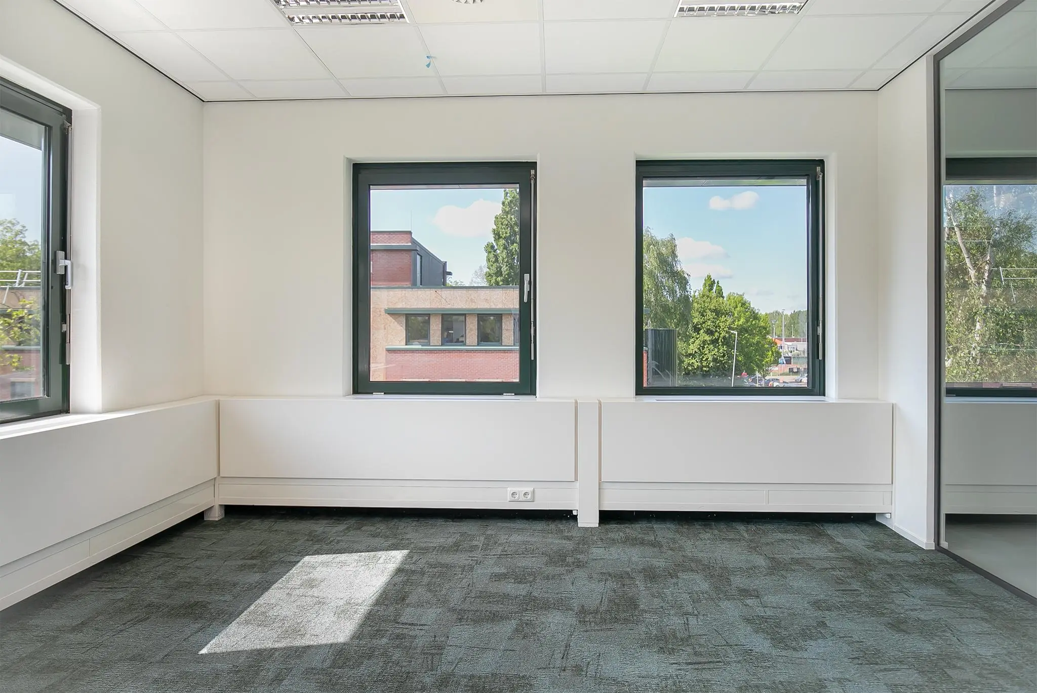 Licht en modern leeg kantoor aan de Kralingseweg met drie ramen en uitzicht op bomen en een bakstenen gebouw.