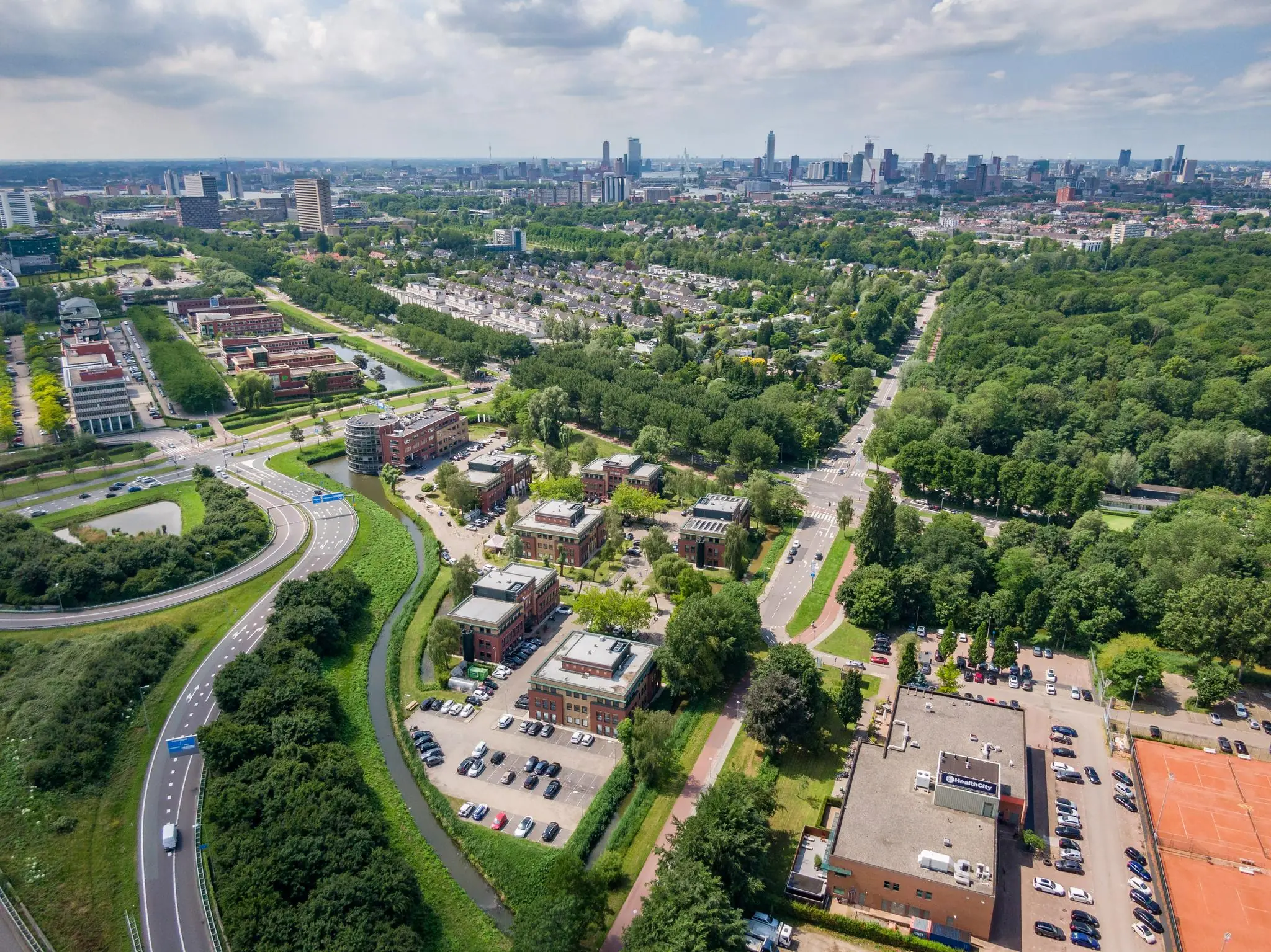 Luchtfoto van de Kralingseweg in Rotterdam met omliggende kantoren, woonwijken, groen en de skyline van de stad op de achtergrond.