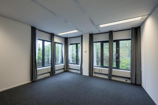 Licht en leeg kantoor met grote ramen, grijze gordijnen en uitzicht op groen.