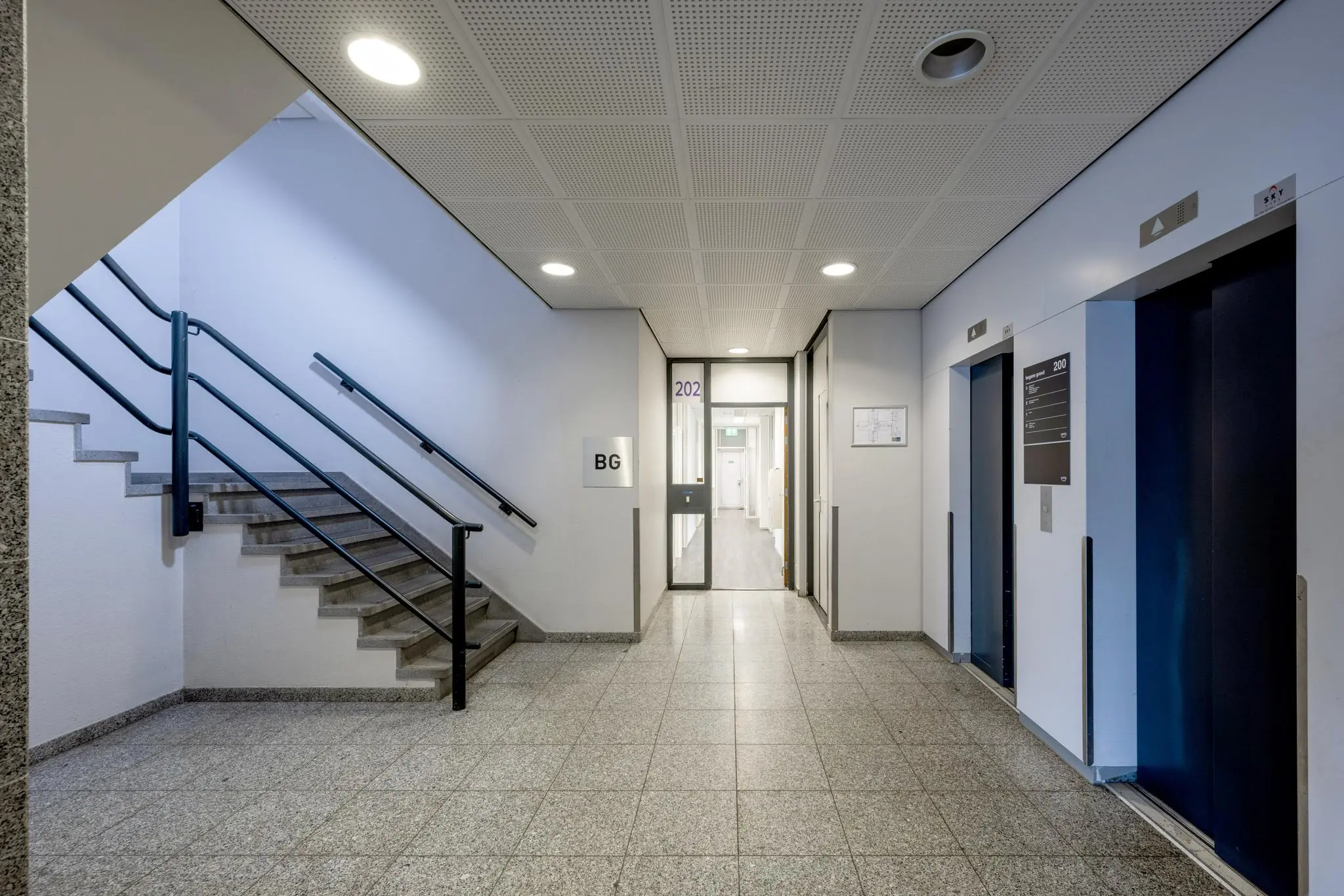 Moderne en nette binnenruimte van een kantoorpand met trap, twee liften en gang naar kamer 202.
