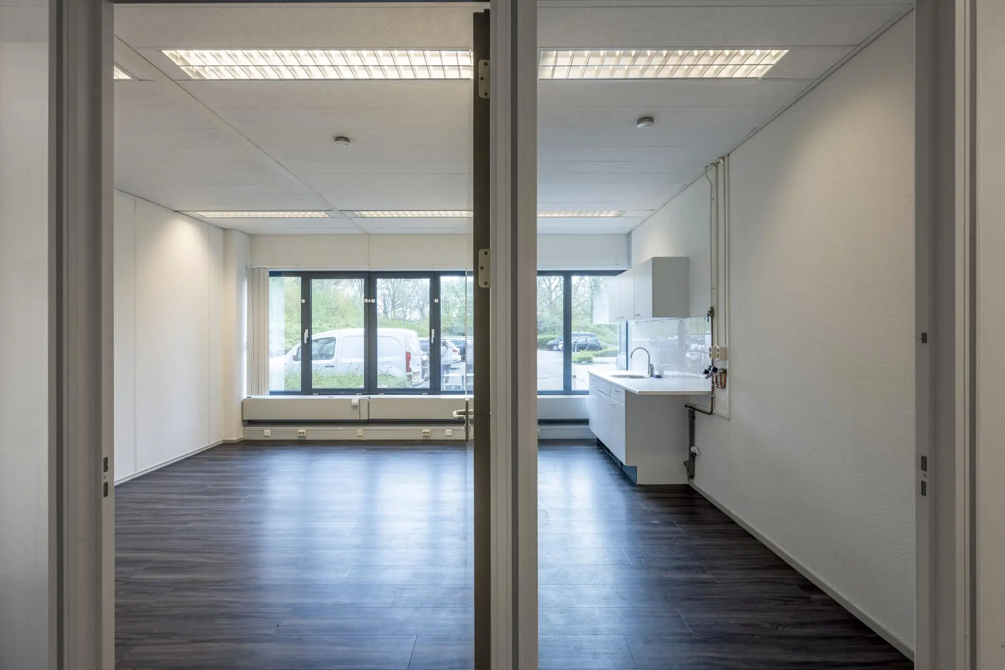 Lege bedrijfsruimte met donker laminaat en een kleine keukenhoek bij grote ramen.