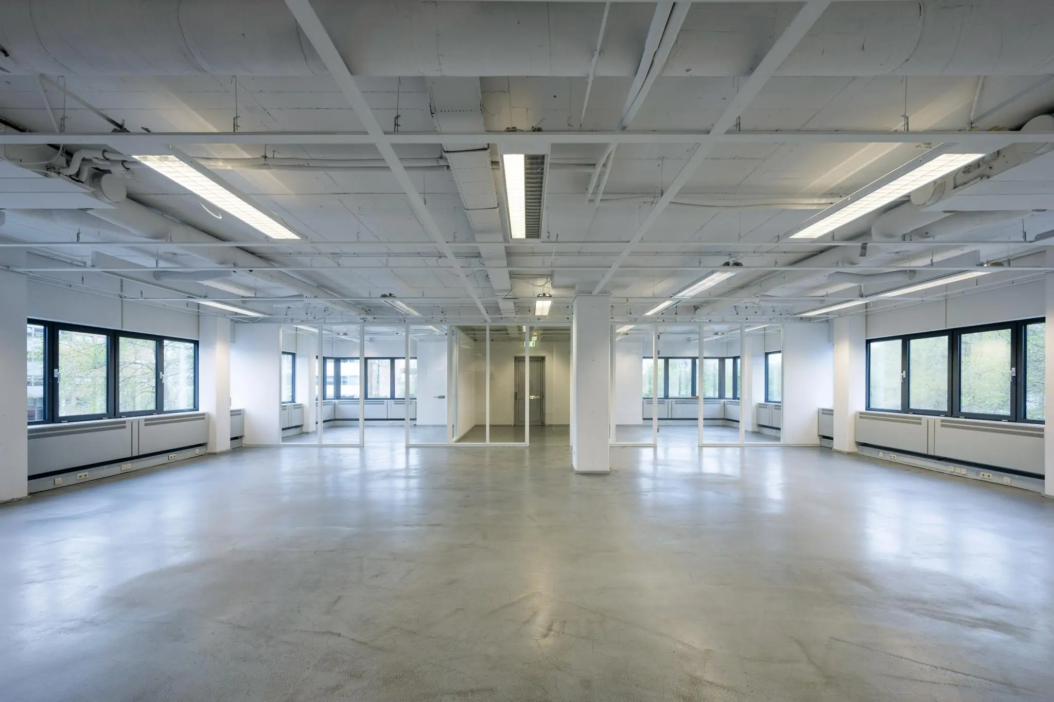 Lichte, lege kantoorruimte met betonnen vloer, zwarte raamkozijnen en glazen wanden rond een centrale lift.