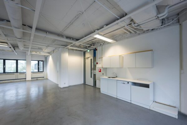 Moderne, lege kantoorruimte met een kleine kitchenette en zicht op grote ramen en een industriële plafondafwerking.