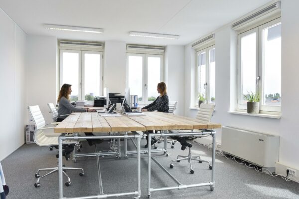 Twee vrouwen werken tegenover elkaar aan een houten vergadertafel in een licht, modern kantoor in Middenburcht.