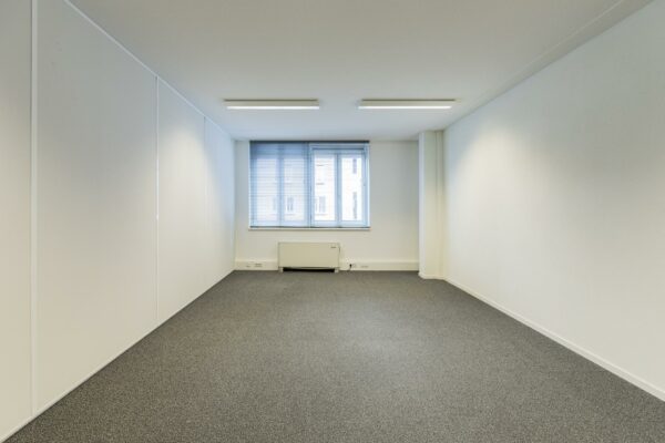 Lege, lichte kantoorruimte in het gebouw Middenburcht met grijs tapijt en een raam met jaloezieën.
