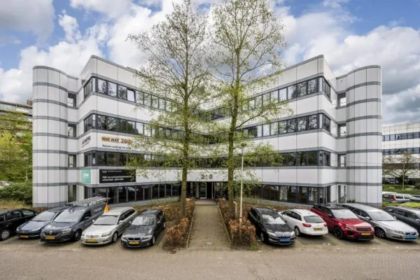 Wit kantoorgebouw met meerdere verdiepingen, parkeergelegenheid en de hoofdingang in het midden, met zichtbare bedrijfsnamen zoals "Your Way 2 Go".