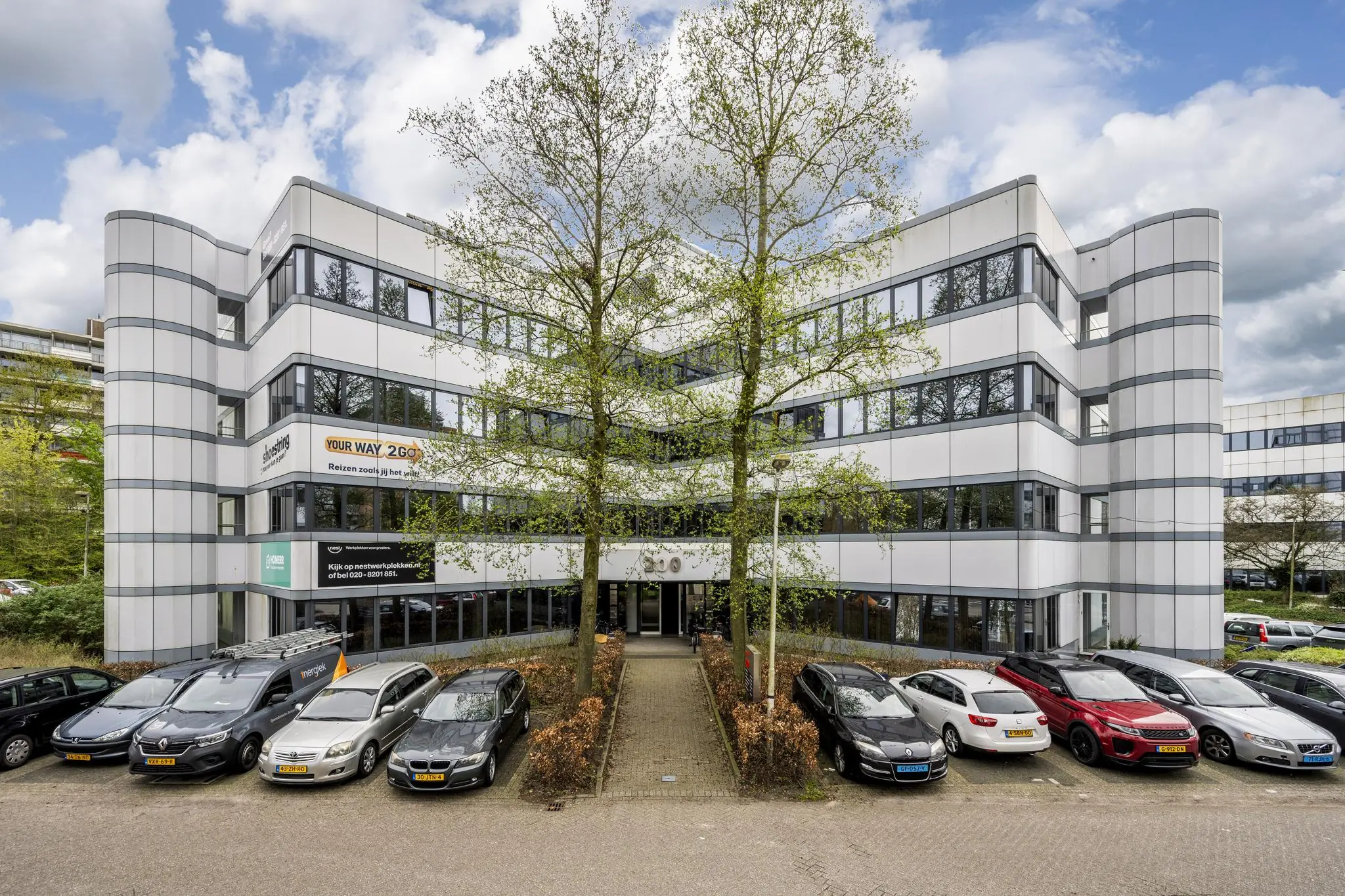 Wit kantoorgebouw met meerdere verdiepingen, parkeergelegenheid en de hoofdingang in het midden, met zichtbare bedrijfsnamen zoals "Your Way 2 Go".