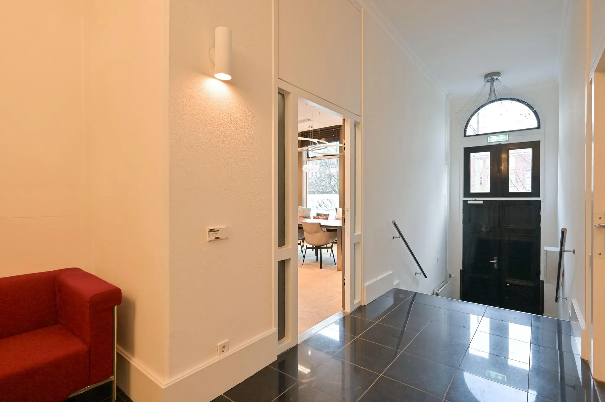 Hal met zwarte tegelvloer, witte muren, een open deur naar een vergaderruimte en een zwarte voordeur met glazen boog.