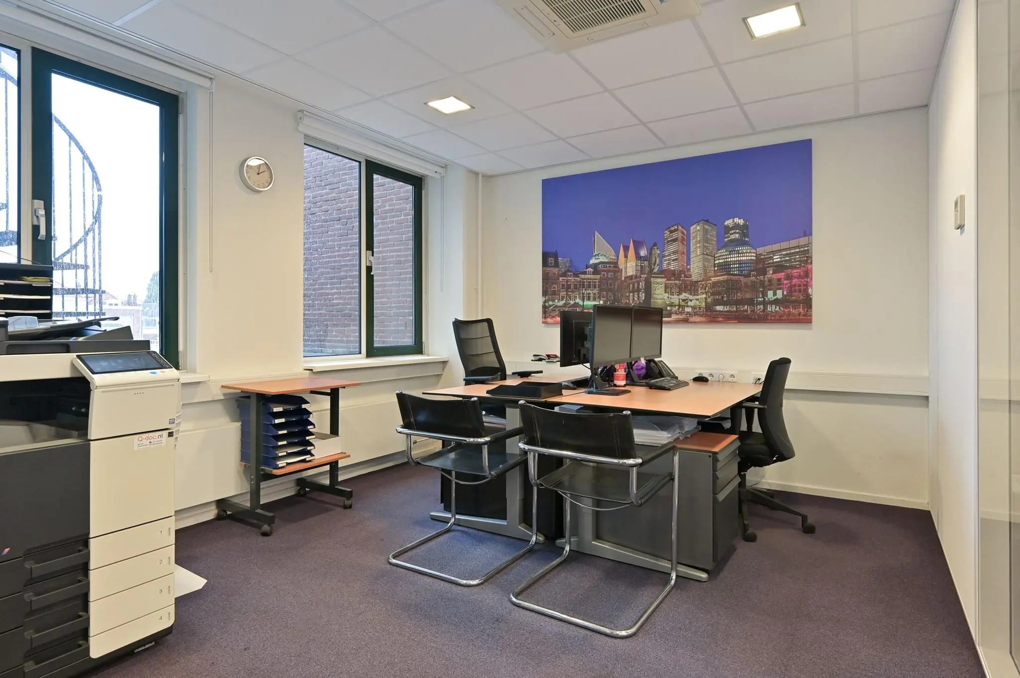 Modern ingericht kantoor aan de Koninginnegracht met bureaus, bureaustoelen, computers, printer en een grote cityscape-muurschildering.