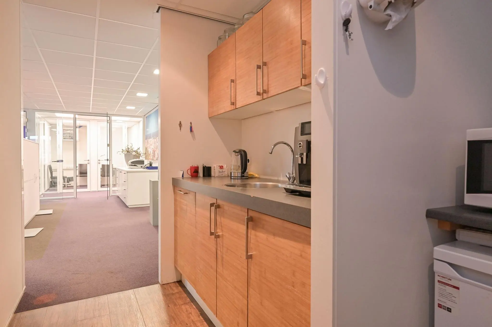 Kleine kantoorpantry met houten keukenkastjes, koffieapparaat en uitzicht op een moderne kantoorruimte aan de Koninginnegracht.