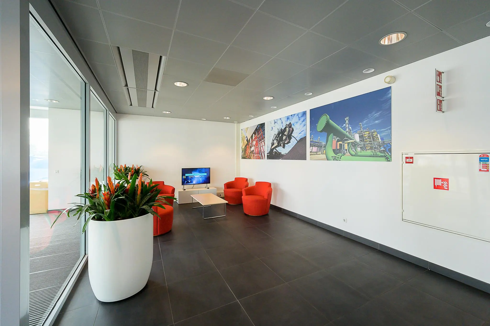 Moderne wachtruimte aan de Willingestraat met rode stoelen, een televisiescherm, planten en industriële foto's aan de muur.