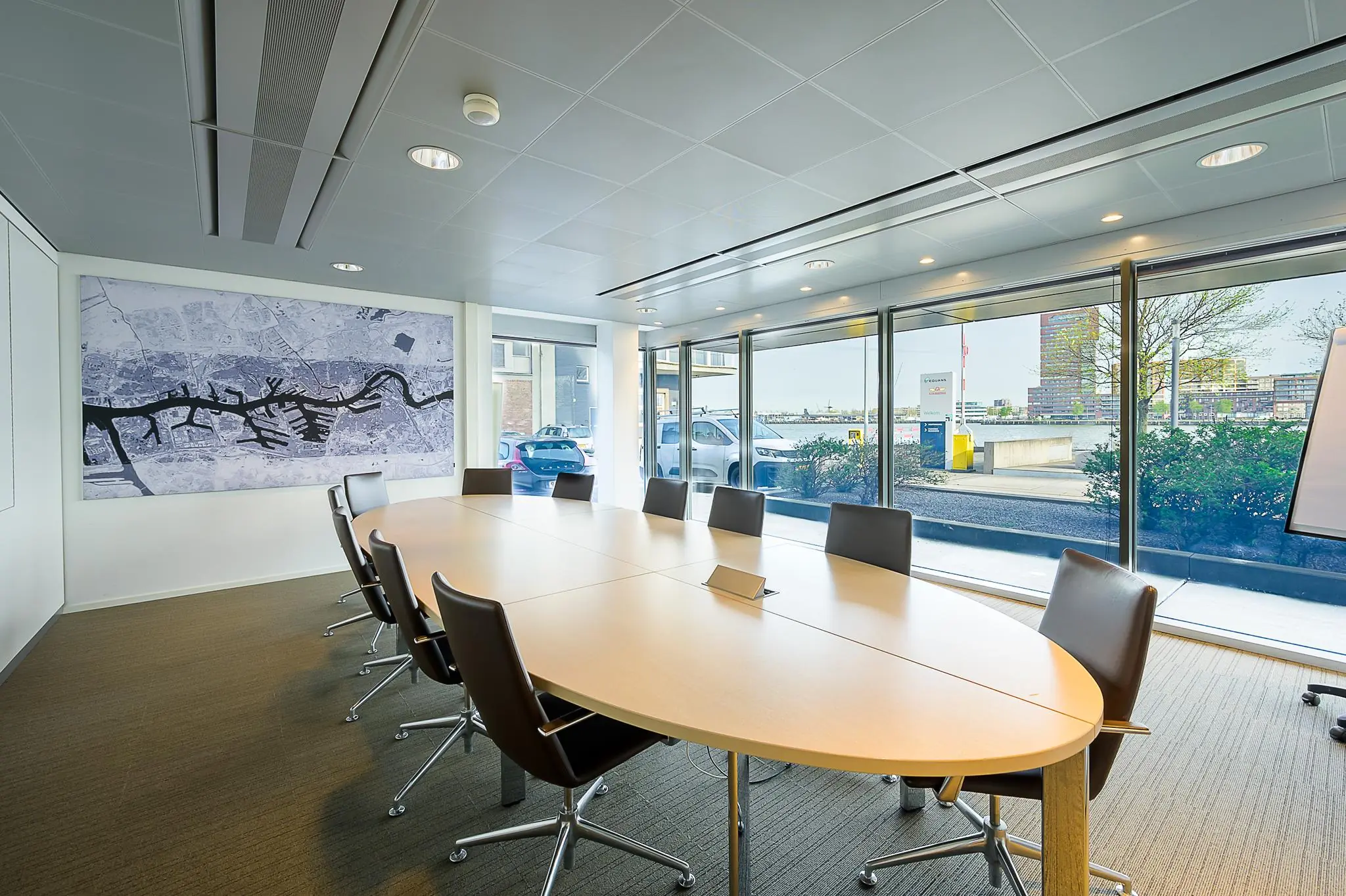 Vergaderruimte aan de Willingestraat met ovale tafel, lederen stoelen, kaart van de stad aan de muur en uitzicht op het water en moderne gebouwen.