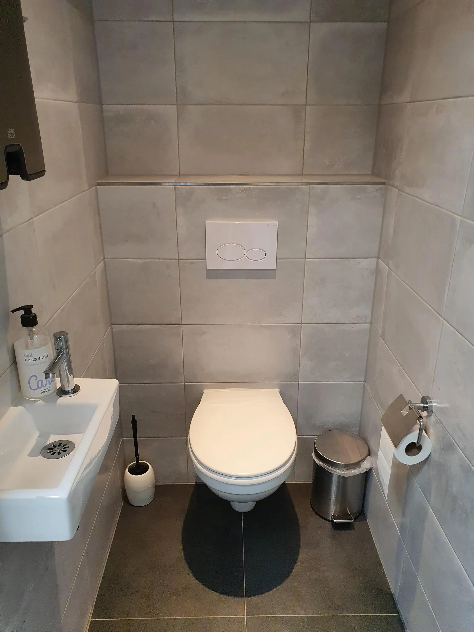 Modern, grijs betegeld toilet met wandcloset, klein fonteintje en accessoires zoals een toiletborstel en prullenbak.