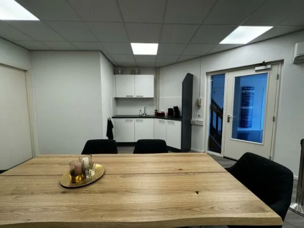 Vergaderruimte met houten tafel, zwarte stoelen, kitchenette en een deur met een 'Bergveste' informatiebord zichtbaar.