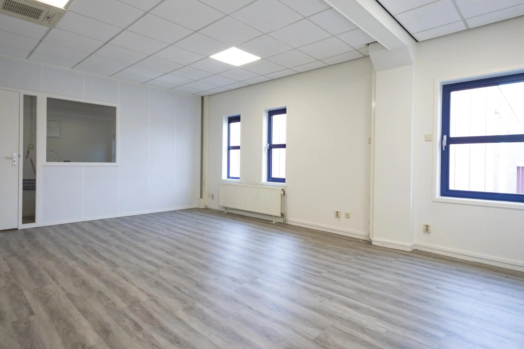 Lichte, lege kantoorruimte met grijze laminaatvloer en drie ramen met donkerblauwe kozijnen in het pand Bergveste.