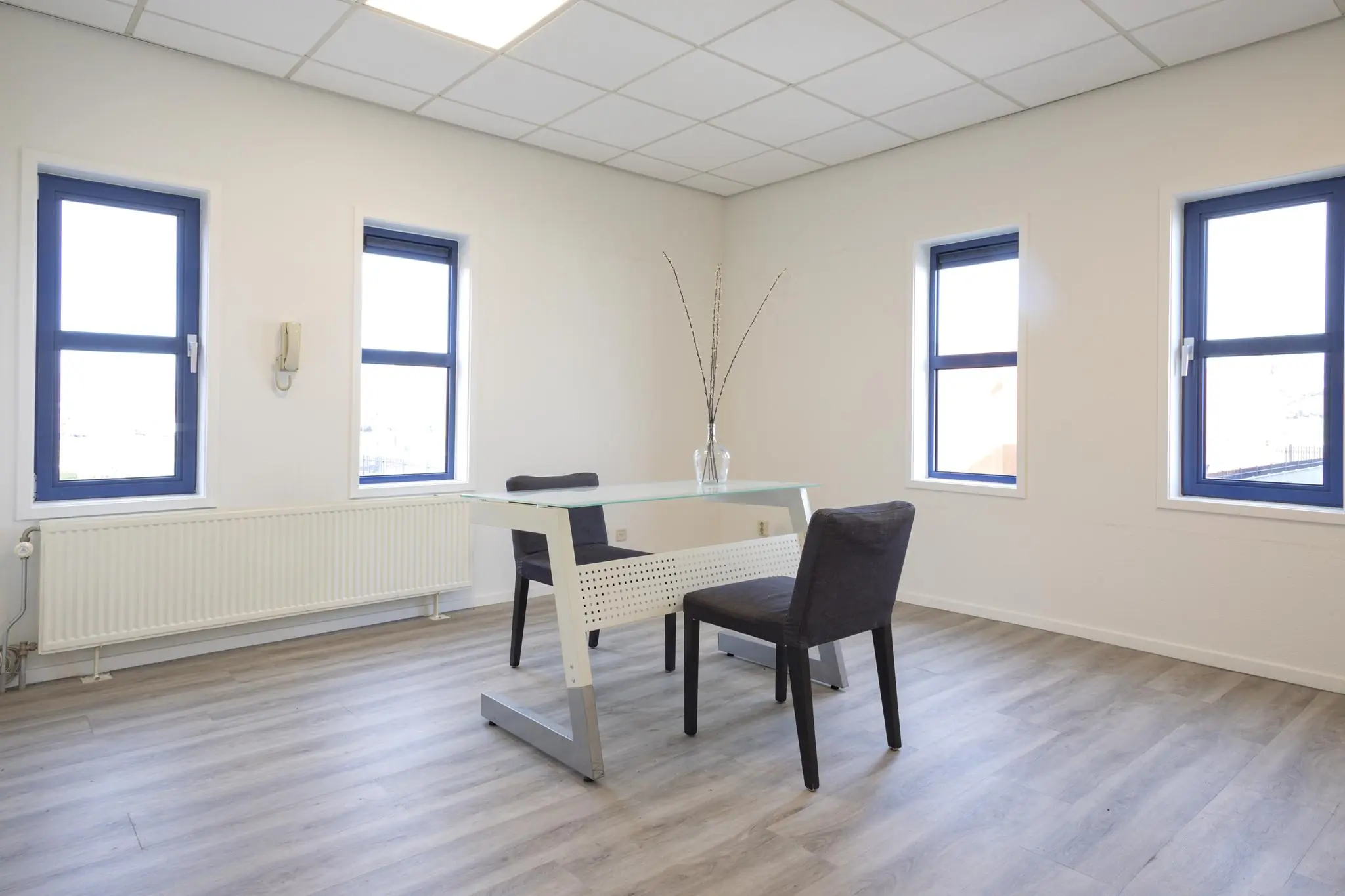 Licht en modern ingericht kantoor met een glazen bureau, twee stoelen en drie ramen met blauwe kozijnen.