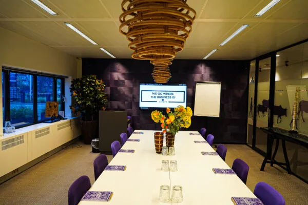 Vergaderruimte aan het Prins Bernhardplein met een lange witte tafel, paarse stoelen, bloemen in het midden en een scherm met de tekst "We go where the business is".