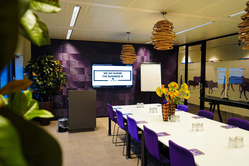 Vergaderruimte op het Prins Bernhardplein met paarse stoelen, een presentatiescherm en bloemen op tafel.