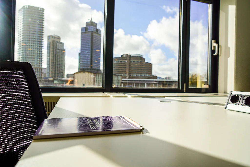Kantoorruimte met uitzicht op het Prins Bernhardplein in Amsterdam, met een notitieboek op een bureau bij het raam.