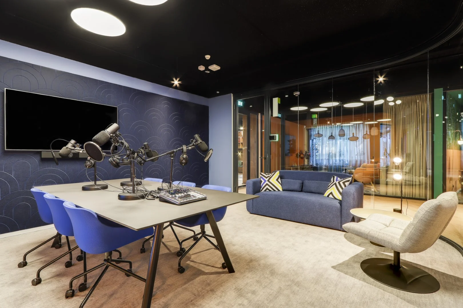 Moderne podcaststudio in Sloterdijk met microfoons, opnametafel, blauwe stoelen en een loungehoek met bank en designstoel.