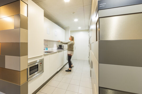 Een vrouw opent een kast in een moderne kantoorpantry met een magnetron en koffiezetapparaat.