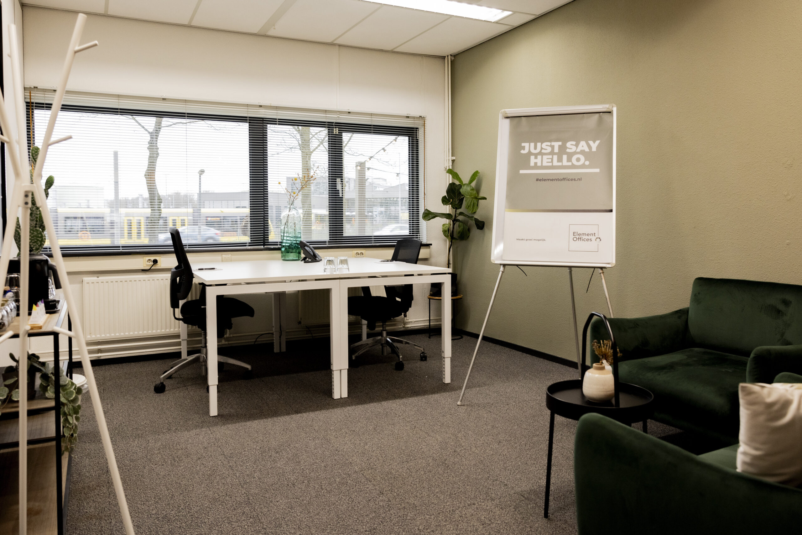 Moderne kantoorruimte met een bureau, stoelen, groene plant, flip-over en bank, en een groot raam met uitzicht naar buiten.