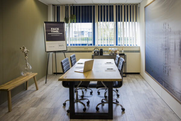 Vergaderruimte met een lange houten tafel, stoelen, een flip-over met de tekst "FACE TIME", en een raam met jaloezieën.