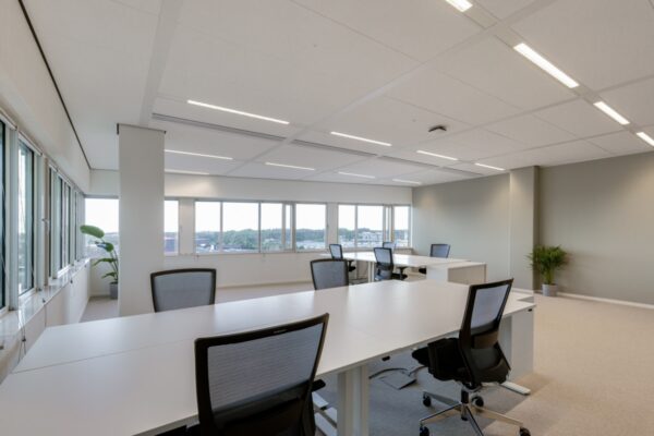 Een modern, licht kantoor met meerdere bureaus en bureaustoelen bij grote ramen.