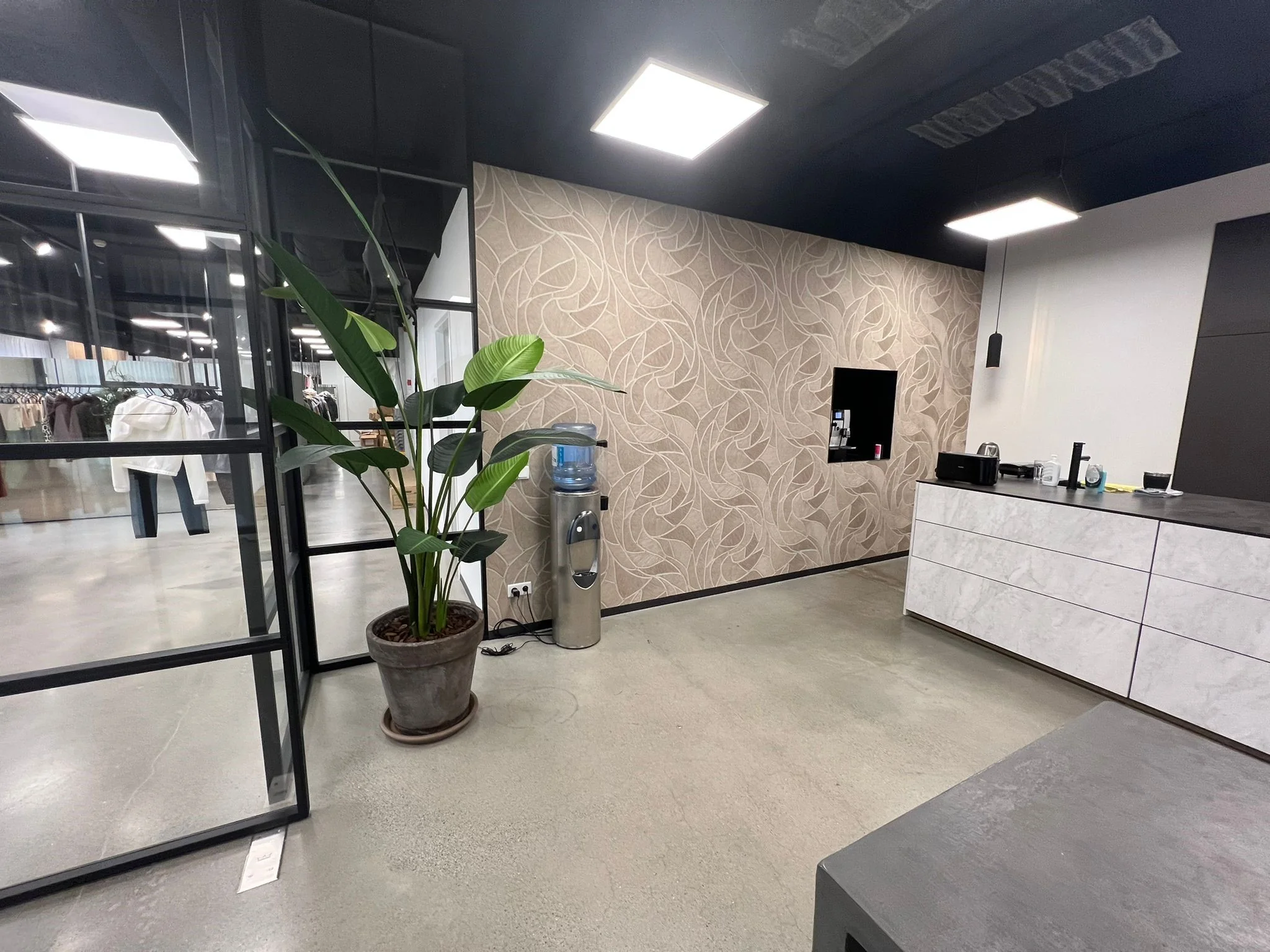 Moderne kantoorruimte aan de Frankfurtstraat met een waterkoeler, grote kamerplant, open keuken en zicht op een showroom met kledingrekken.