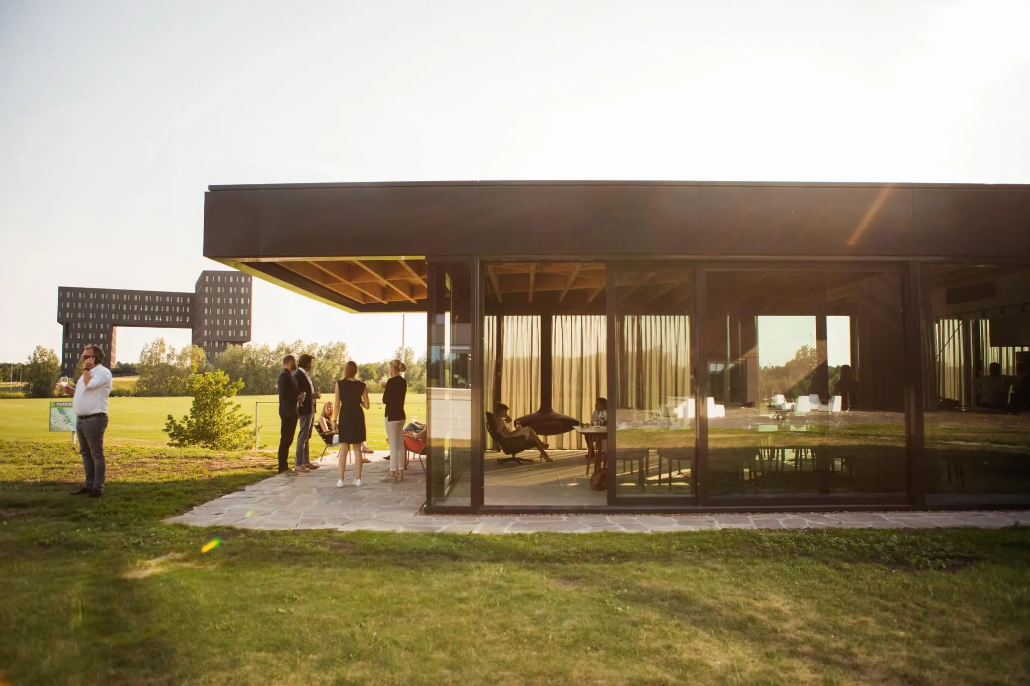 Mensen staan en zitten buiten bij een modern glazen paviljoen aan de Orteliuslaan met een markant zwart gebouw op de achtergrond.