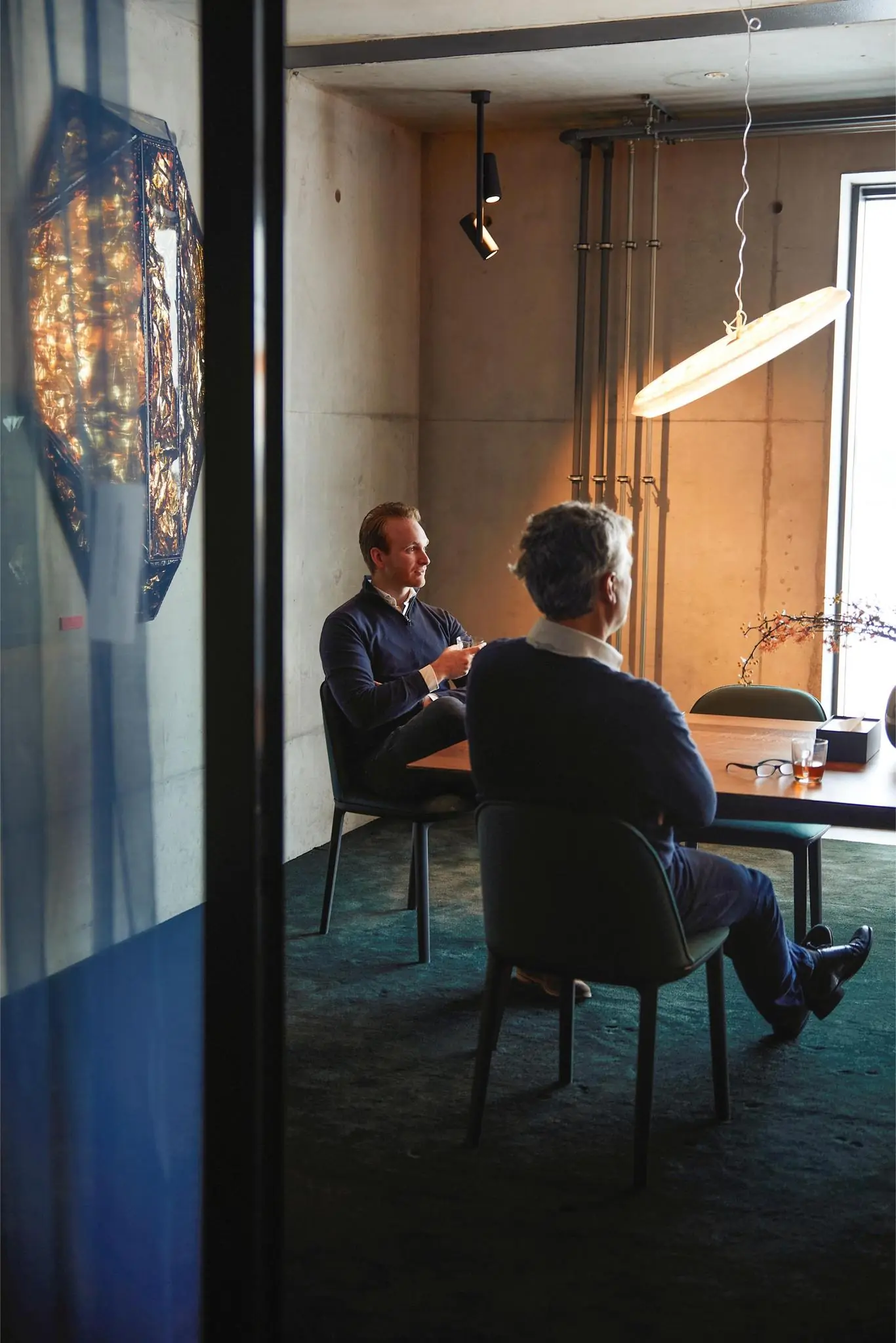 Twee mannen zitten in een modern vergaderkamerinterieur aan de Orteliuslaan, met warm licht en industriële elementen.